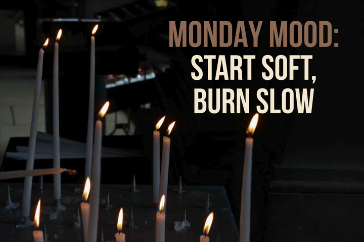 Lit candles in a dark room, with text that reads: “Monday Mood: Start soft, burn slow.”